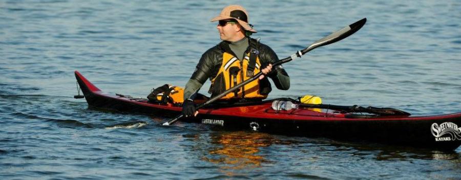 Jeff Fabiszewski is sitting in a red and black sea kayak on calm water. He is wearing a tan wide-brimmed hat, dark sunglasses, a long-sleeve paddle jacket, gloves, and an orange life jacket. He is holding a double-bladed paddle across the kayak, with one blade in the water. On the deck of the kayak are a yellow dry bag, water bottles, and other small gear secured with bungee cords. The sea kayak is a Nigel Dennis NDK Greenlander Pro, designed by British Sea Kayaks. The background shows open water with other paddlers and boats far in the distance.