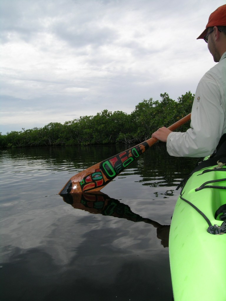 Jeff Fabiszewski uses a wooden canoe paddle with a painted raven blade beside mangroves, its reflection visible alongside a green kayak.