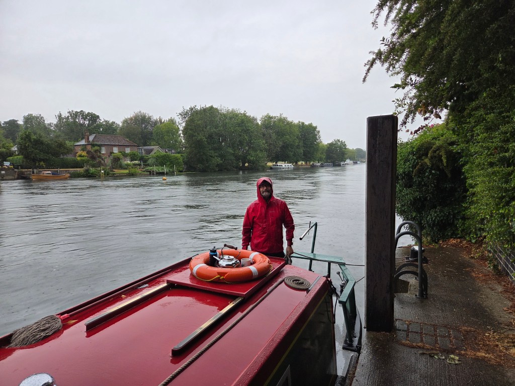 On the stern deck of a narrowboat, you stand steady in the rain, rope in hand and line wrapped around a bollard, keeping the vessel secure on a wet Thames morning. The damp sheen on the deck and the misty air speak to the realities of river travel where rain is as constant a companion as current and tide. With your practiced stance and calm presence, the moment reflects the quiet discipline of seamanship: tending lines, braving weather, and carrying on the timeless work of the waterways.