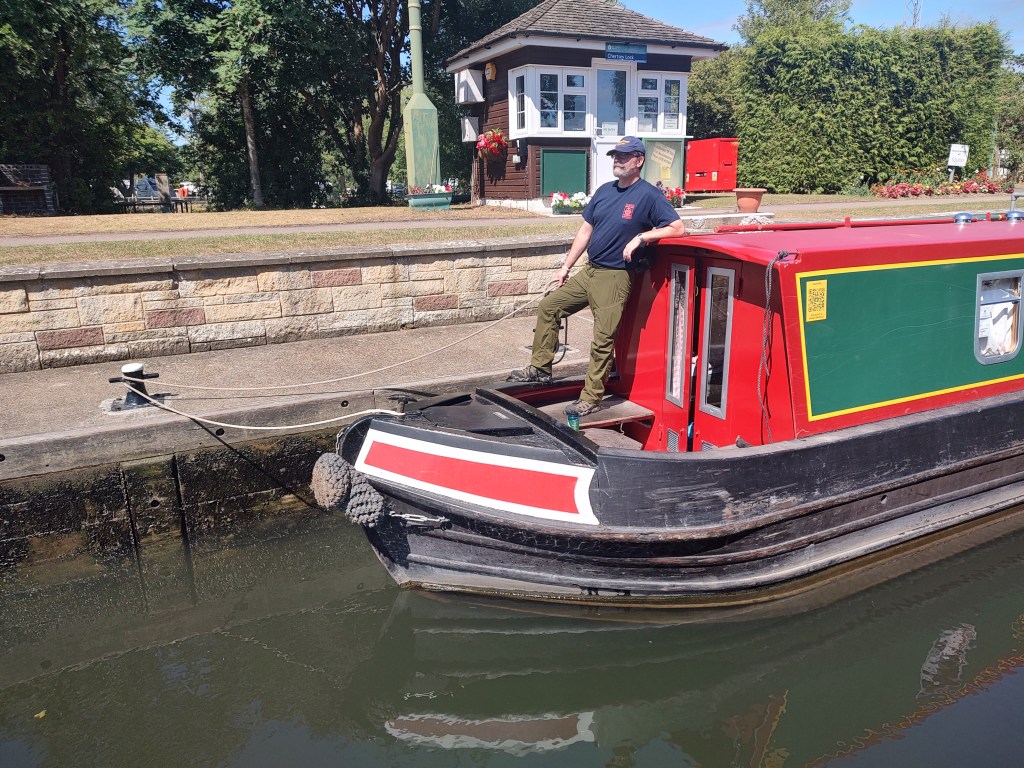 On the bow deck of a red and green narrowboat, you stand confidently with a line secured around a bollard, guiding the vessel through one of the River Thames’ many historic locks. Dressed in a navy shirt, olive trousers, and your Sea Scout cap, you embody both seamanship and tradition. The lock-keeper’s house and flower baskets in the background add to the timeless charm of the English waterways. This photograph captures the practical art of boat handling, where patience, rope work, and river rhythm come together in a scene as old as canal travel itself.
