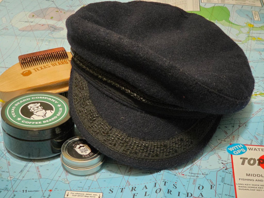 The image shows a small collection of grooming tools and a traditional sailor’s cap resting on a nautical chart. In the center lies a round black tin labeled “Texas Beard Company Coffee Beard Balm.” Next to it is a smaller silver tin of mustache wax from the same brand. A wooden beard comb stamped with the Texas Beard Company logo leans against the balm, with a natural-bristle beard brush placed nearby. Draped across the items is a navy wool Breton cap, the kind traditionally worn by English boatmen. The whole arrangement sits on a colorful nautical map of Florida waters, giving the sense of a mariner’s kit laid ready for voyage.