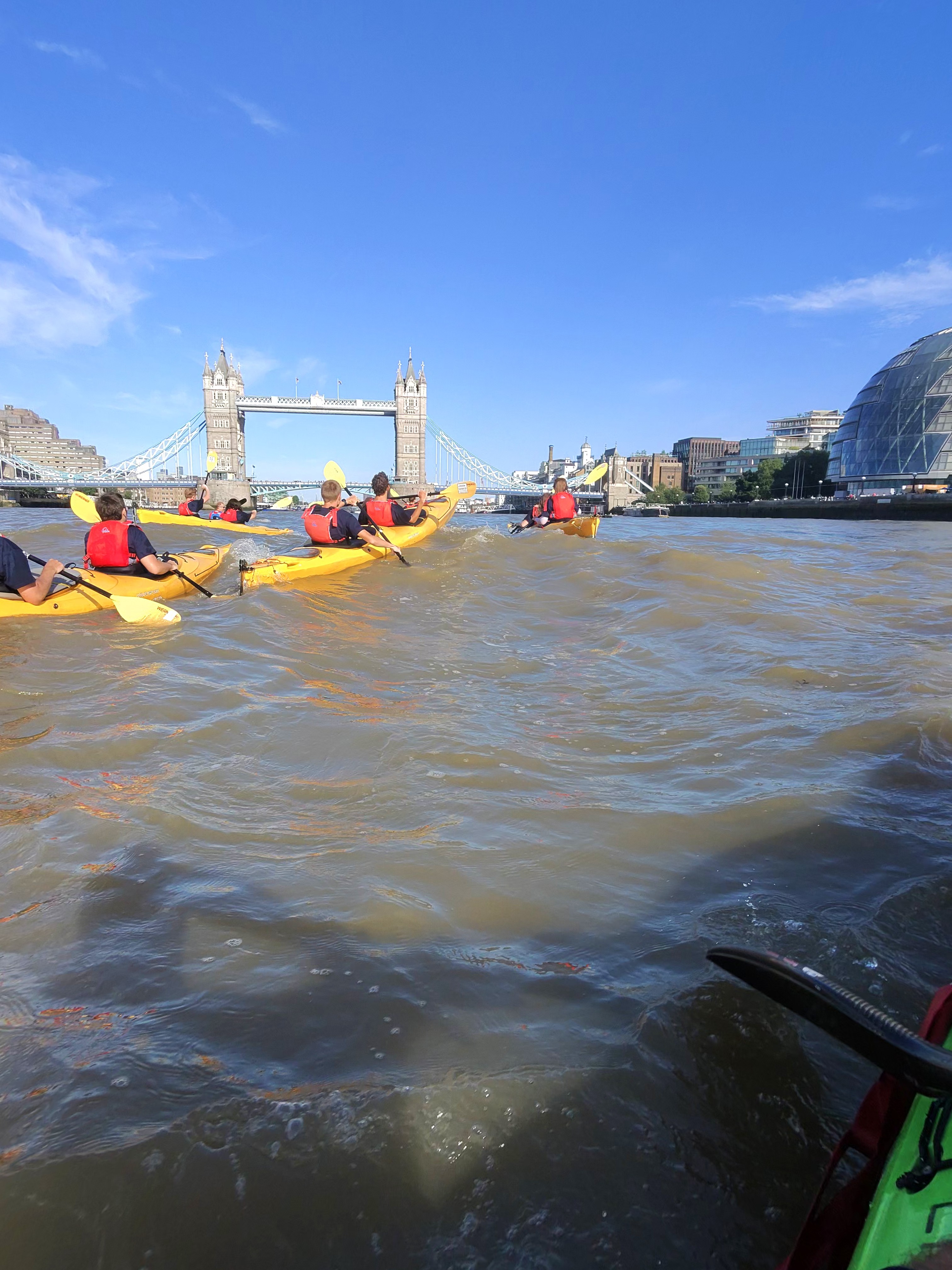 Group of kayakers wearing red life jackets paddling yellow kayaks on the tidal River Thames toward Tower Bridge in London.