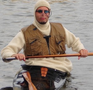 Jeff Fabiszewski wears a hooded tuilik and tan life jacket while holding a wooden Greenland paddle across a black kayak