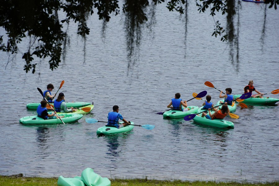 Group of young paddlers wearing life jackets practicing kayak handling in green recreational kayaks on calm water.