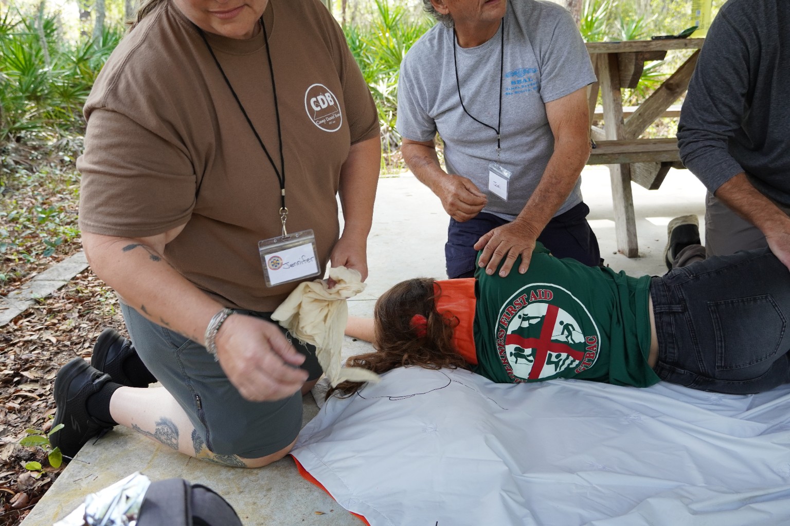 Wilderness First Aid participants kneel beside a simulated patient lying on their side on a groundsheet beneath an outdoor pavilion.